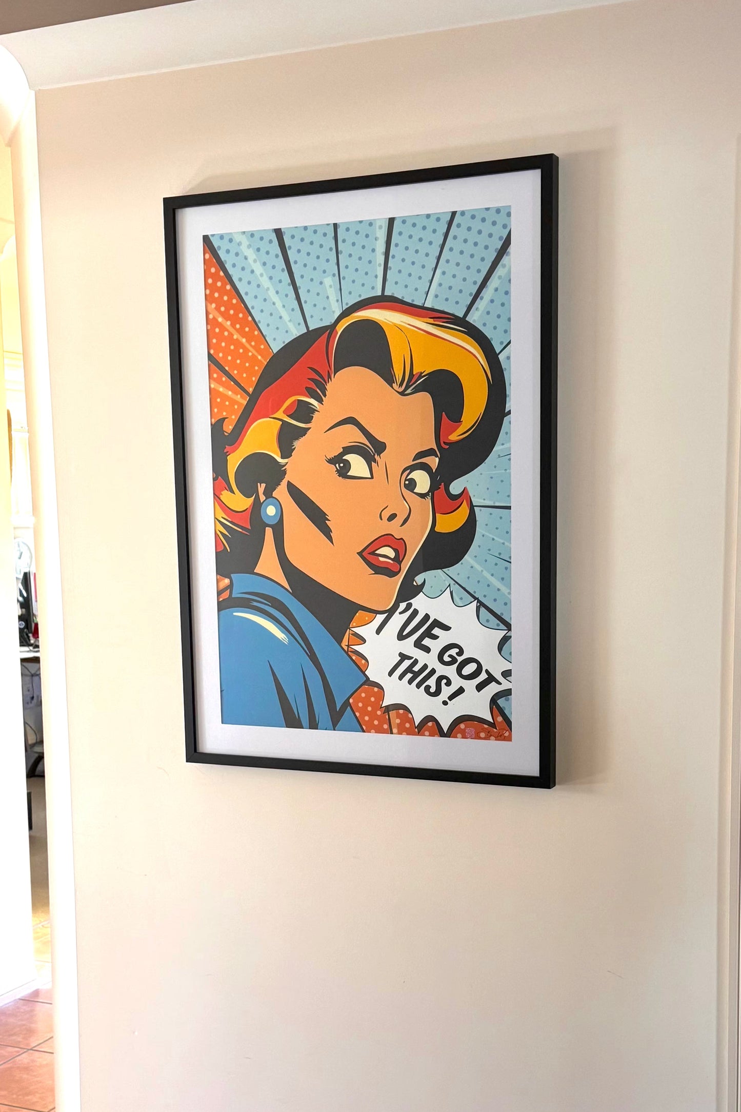 A framed pop-art poster featuring a retro comic-style woman with the text 'I've got this' hanging on a white wall in an indoor hallway with tiled floor.