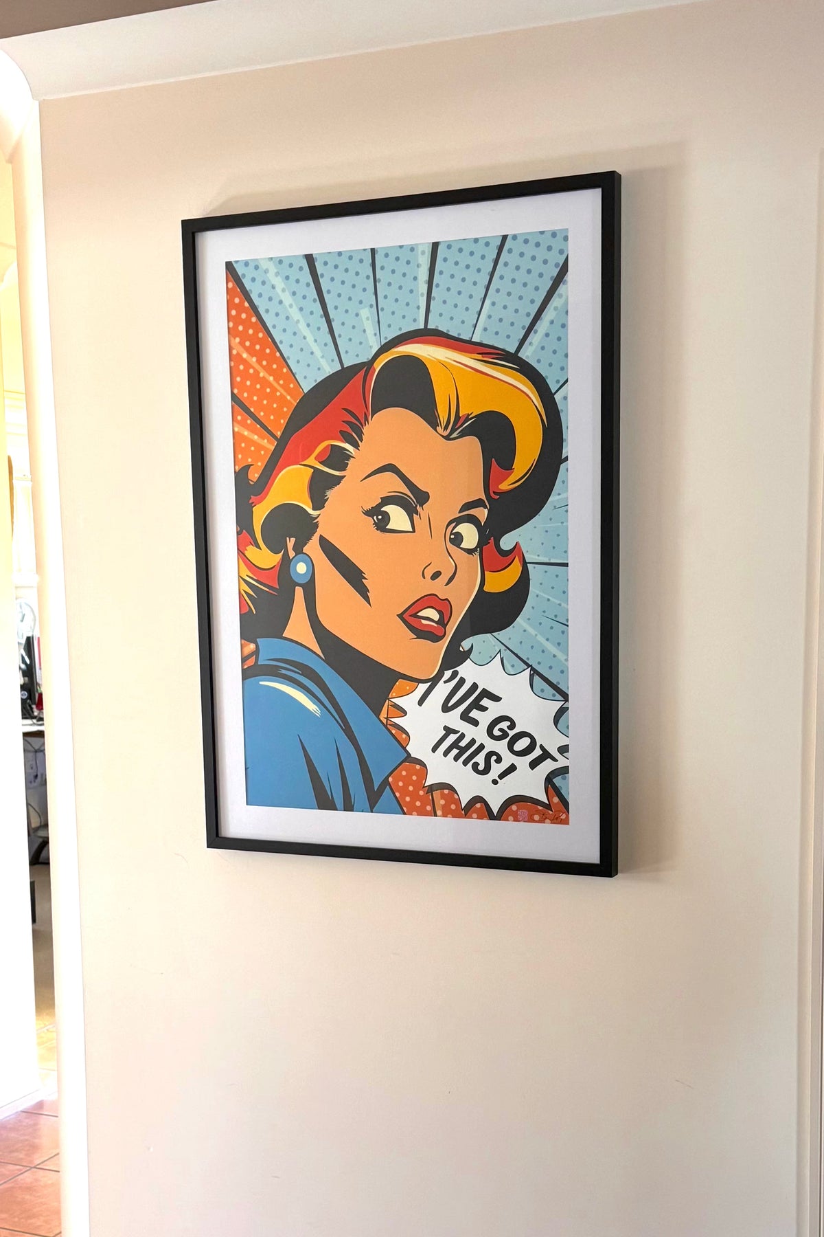 A framed pop-art poster featuring a retro comic-style woman with the text 'I've got this' hanging on a white wall in an indoor hallway with tiled floor.